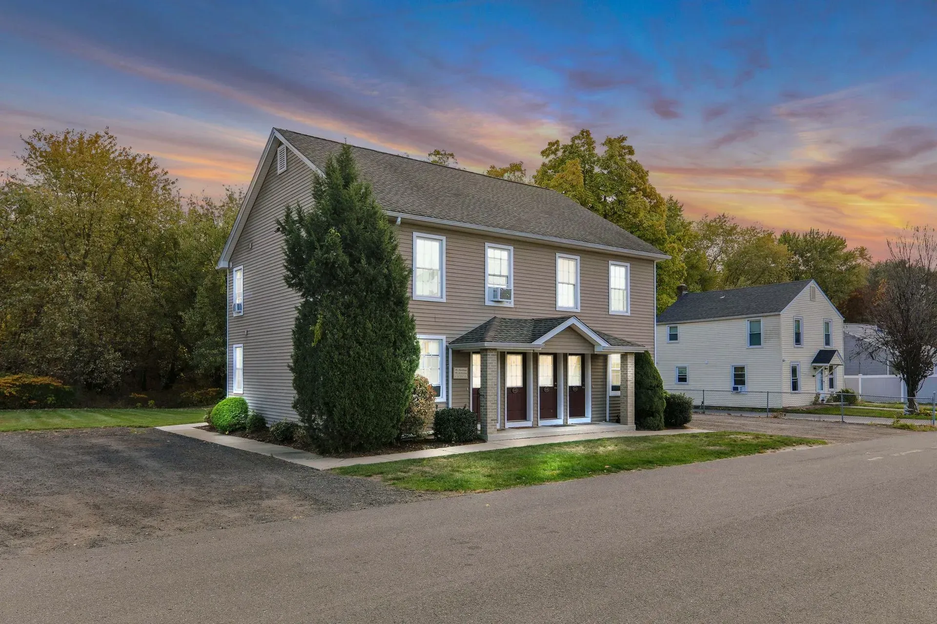 A gray two-story house with a front porch at dusk, set beside a gravel road and trees.