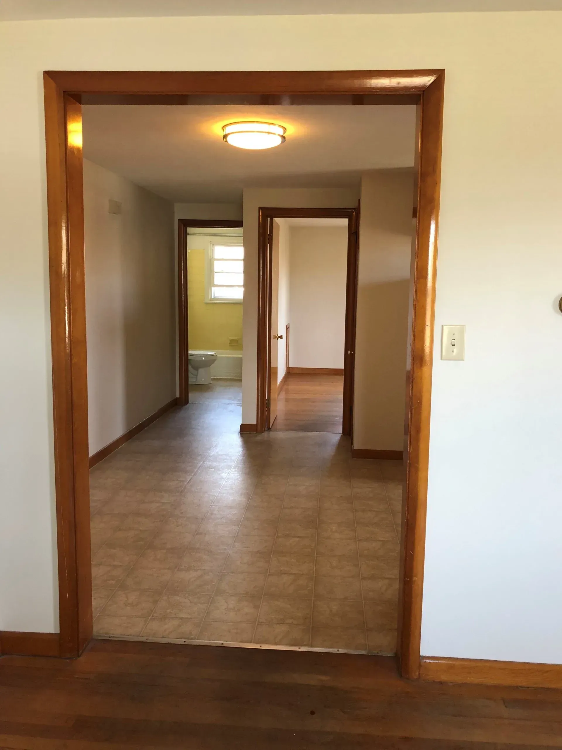 Interior hallway with wood-trimmed doorway, tiled floor, and a lit ceiling fixture leading to rooms ahead