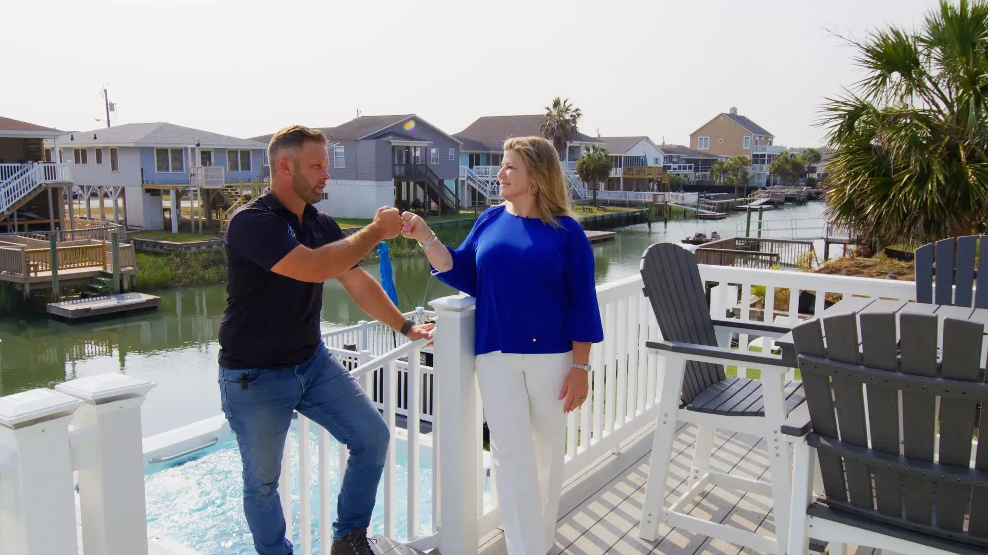Two people talking on a white waterfront deck with houses and boats in the background.