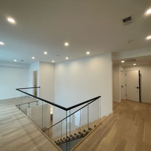 Bright modern hallway with wood floors, white walls, recessed lights, and glass stair railing.