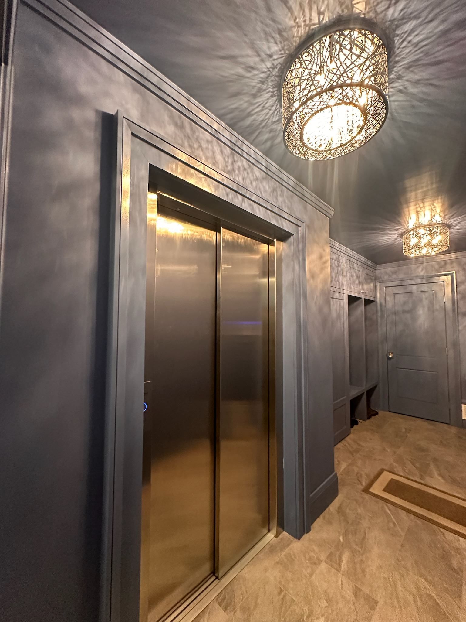 A hallway with a stainless steel elevator and a chandelier hanging from the ceiling.