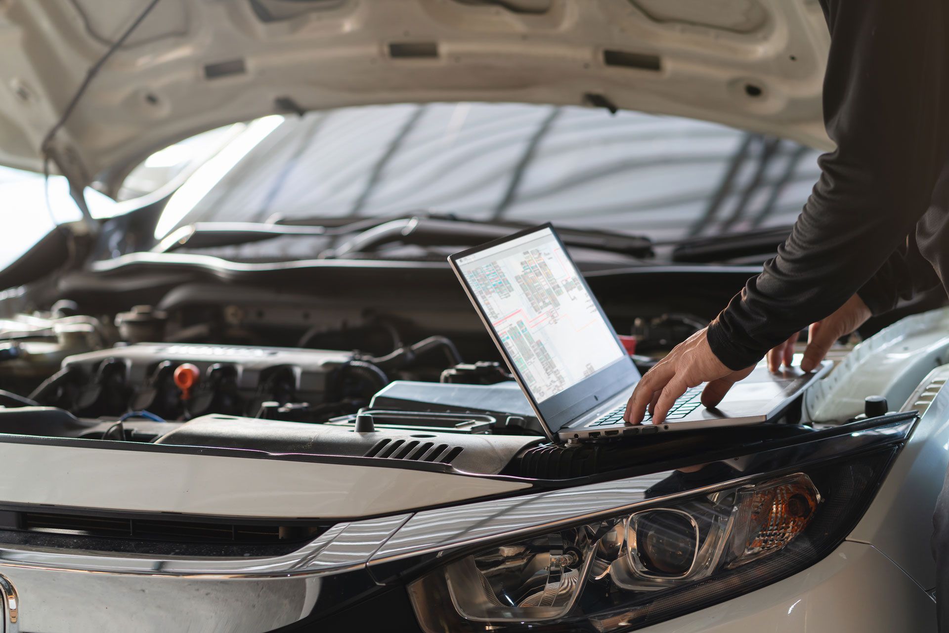 Mechanic uses laptop to diagnose car engine with hood open in a garage.