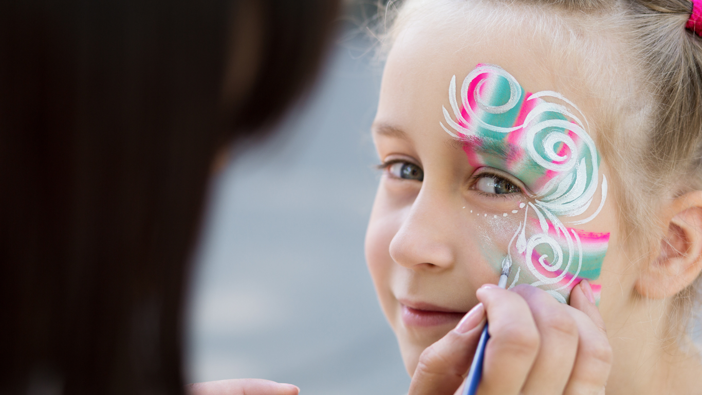 Girl getting face painted with pink, green, and white swirls, smiling.