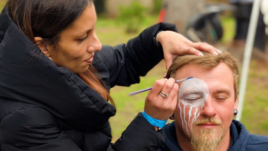 A makeup artist applies white face paint to a man's face outdoors.