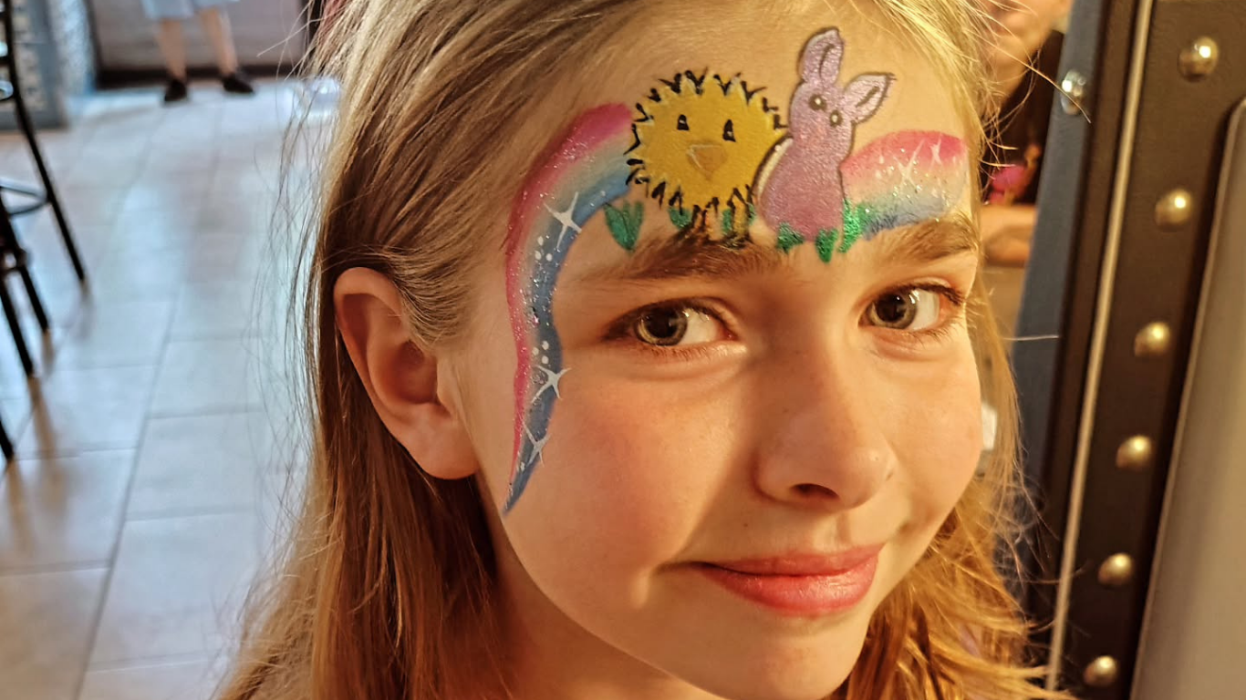 Girl with face painted as a sun, rainbow, and bunny smiling at camera.