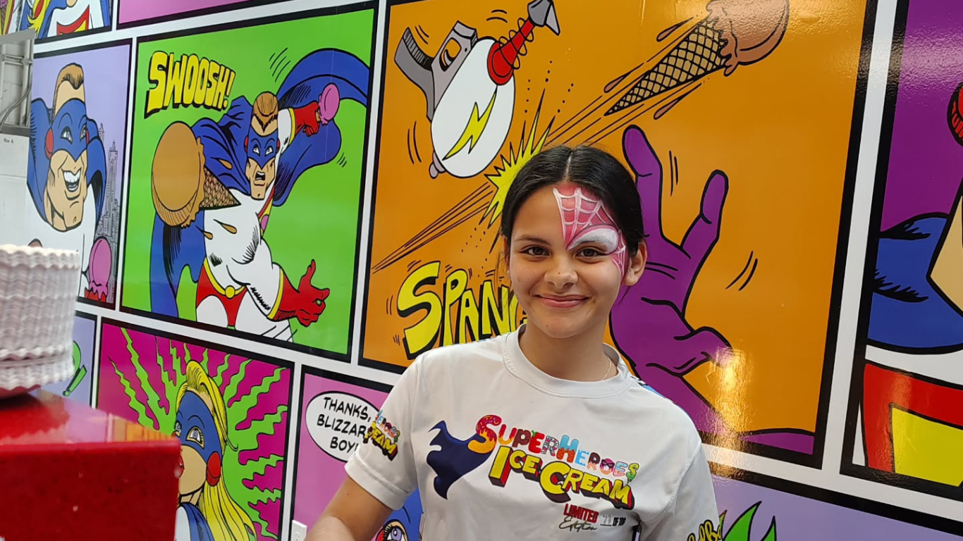 Young person with face paint, smiling, in front of superhero-themed wall.
