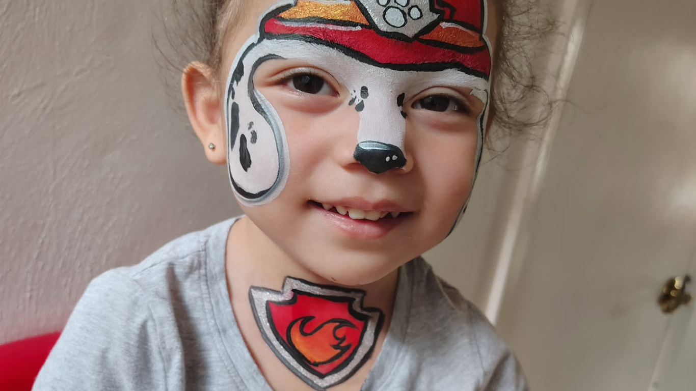 Child with Marshall the Paw Patrol face paint, smiling. The child also has a Paw Patrol logo on the neck.