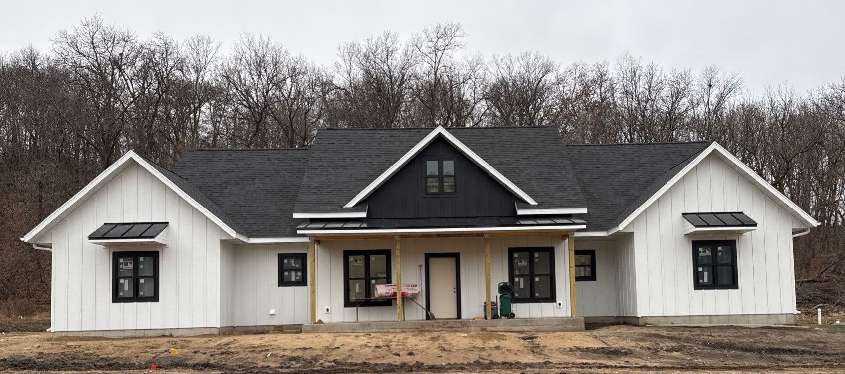 White farmhouse under construction with black accents and roof, set against a backdrop of bare trees.