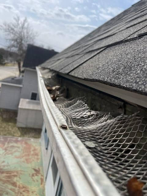 Gutter filled with debris, covered by mesh screen, on a house roof. Cloudy sky in the background.