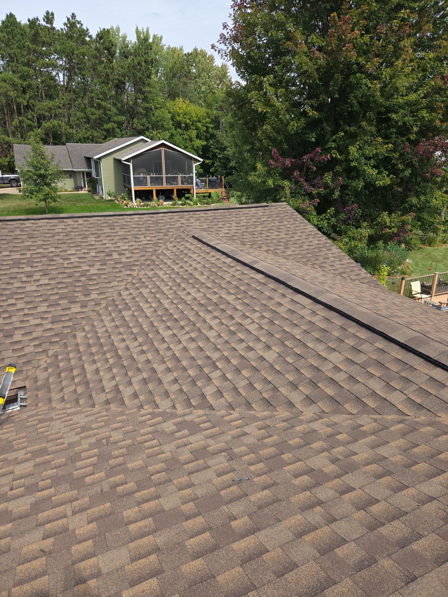 Brown shingled roof with a black line along the ridge, green trees, and a house in the background.