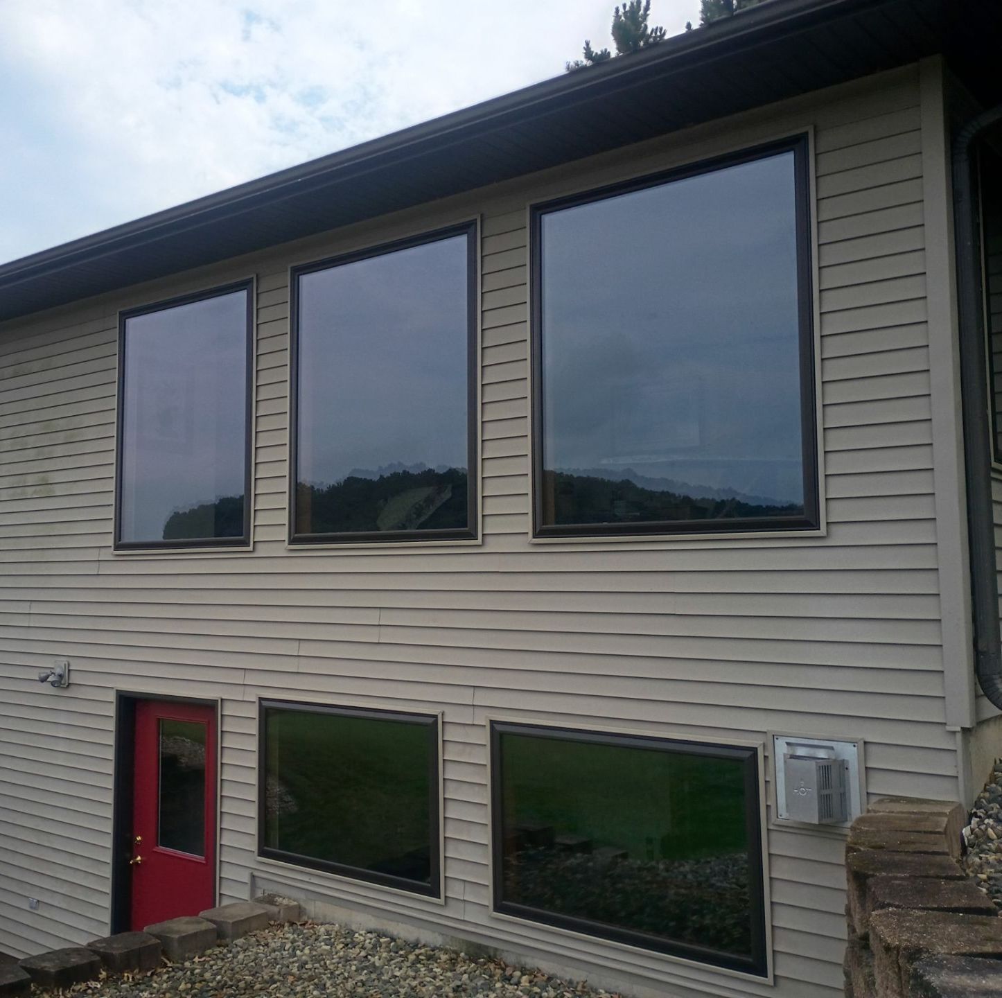 Beige house with three large, dark-tinted windows above two smaller windows. A red door is on the side.
