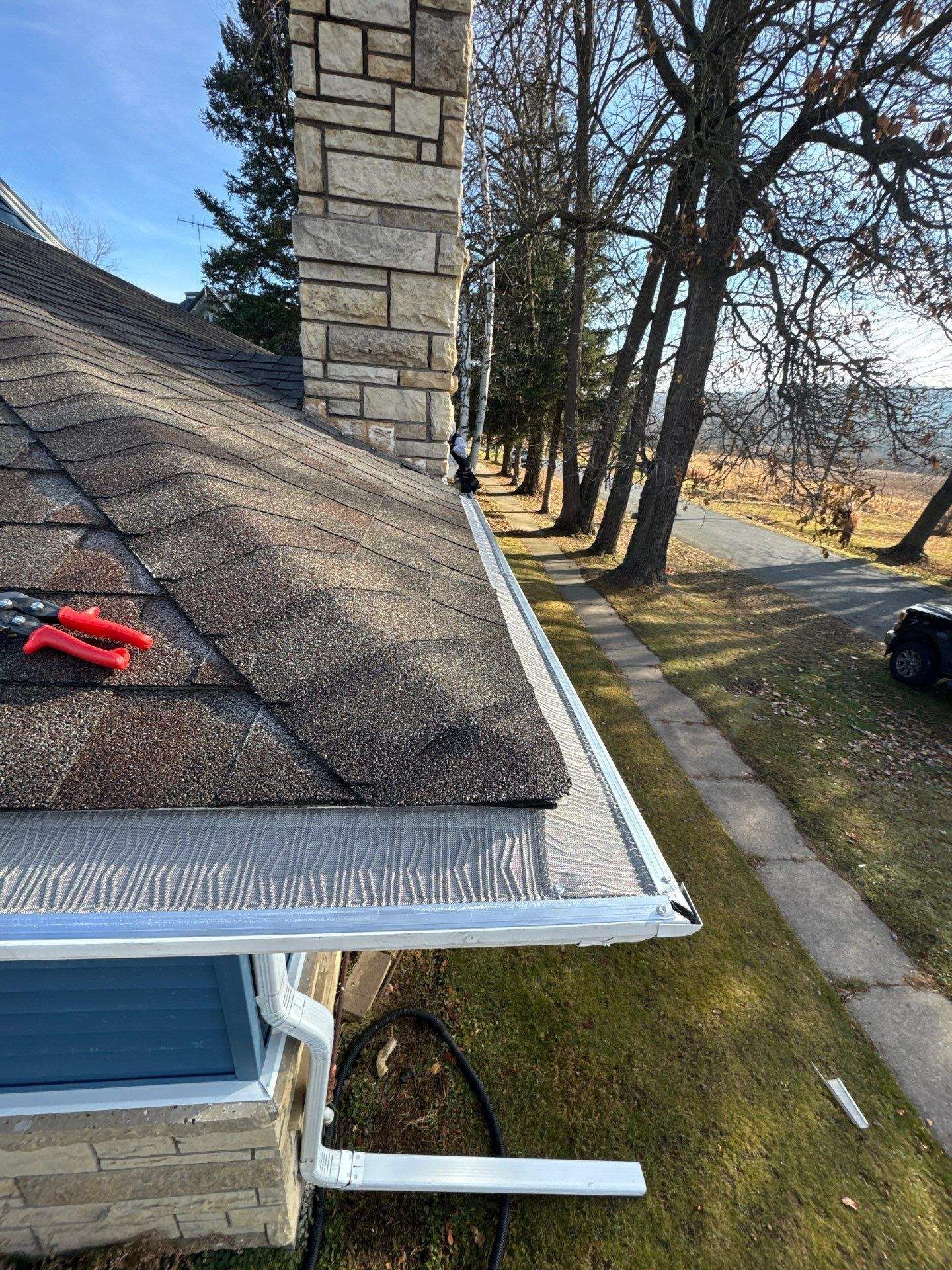 View of a roof with a newly installed gutter. Chimney and trees in the background. Blue sky.