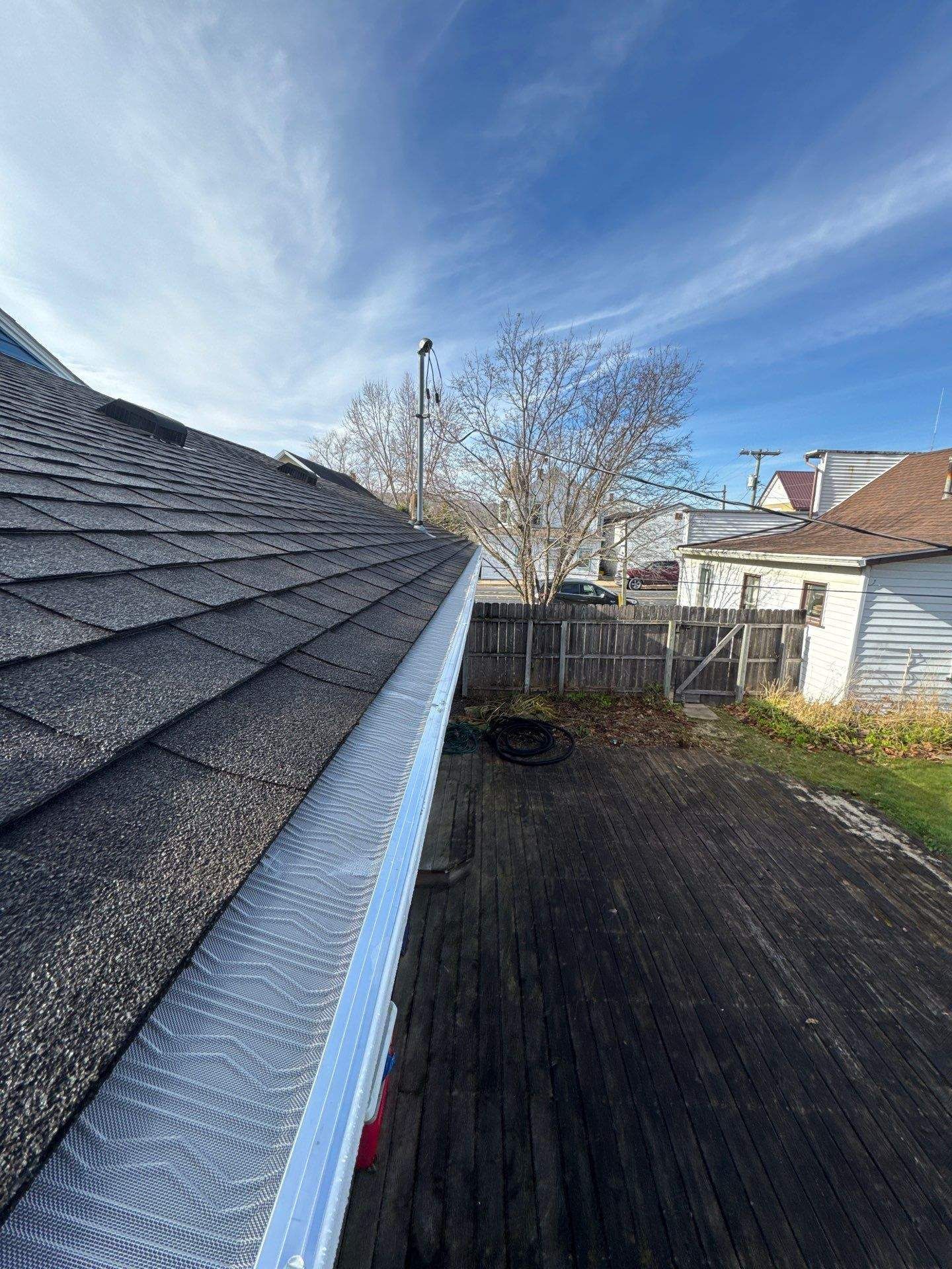 Roof and gutter with wooden deck and fence in background under blue sky.