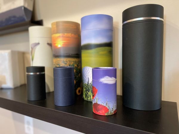 Several cremation urns of various sizes and designs on a dark shelf, including ones with sunset, landscape, and floral patterns.