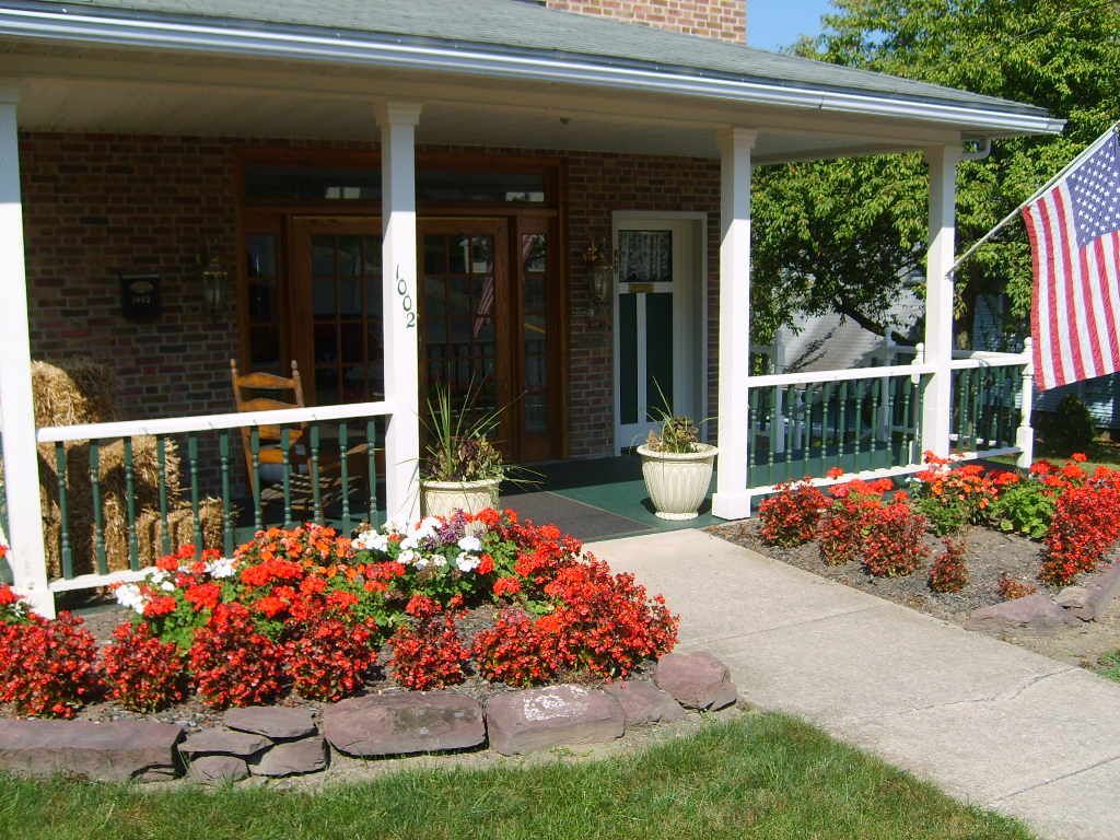 Red flowers and stone border a walkway to a brick house with a porch. American flag.