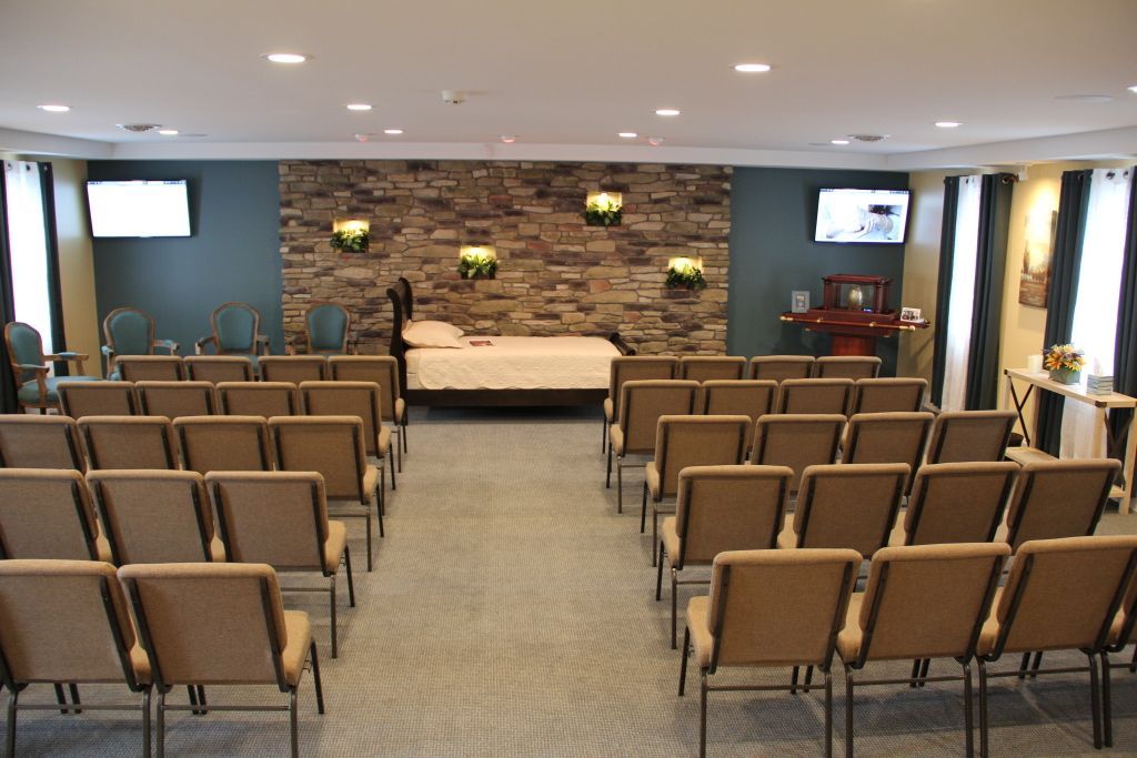 Funeral home chapel with rows of chairs facing a casket on a raised platform, decorated with a stone accent wall and two televisions.