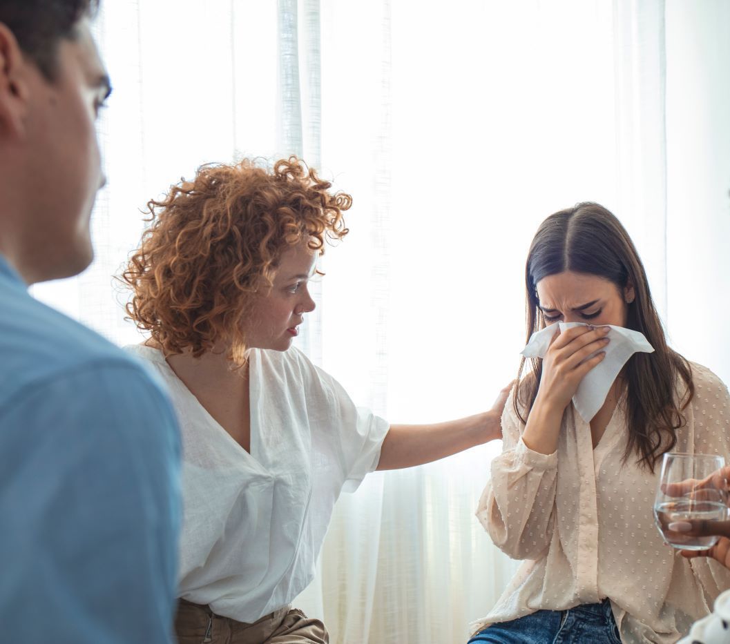 Group therapy session: A woman cries, wiping her face while others offer support; setting is a bright room.