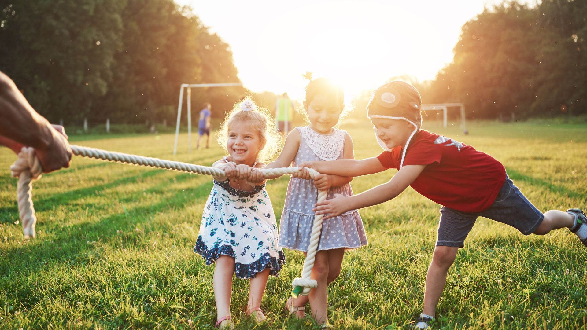 Young children in a park tugging on a rope. Sunset over trees.