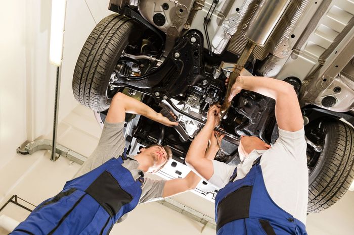 Due meccanici lavorano sotto un'auto su un ponte sollevatore in un'officina, ispezionandola o riparandola.