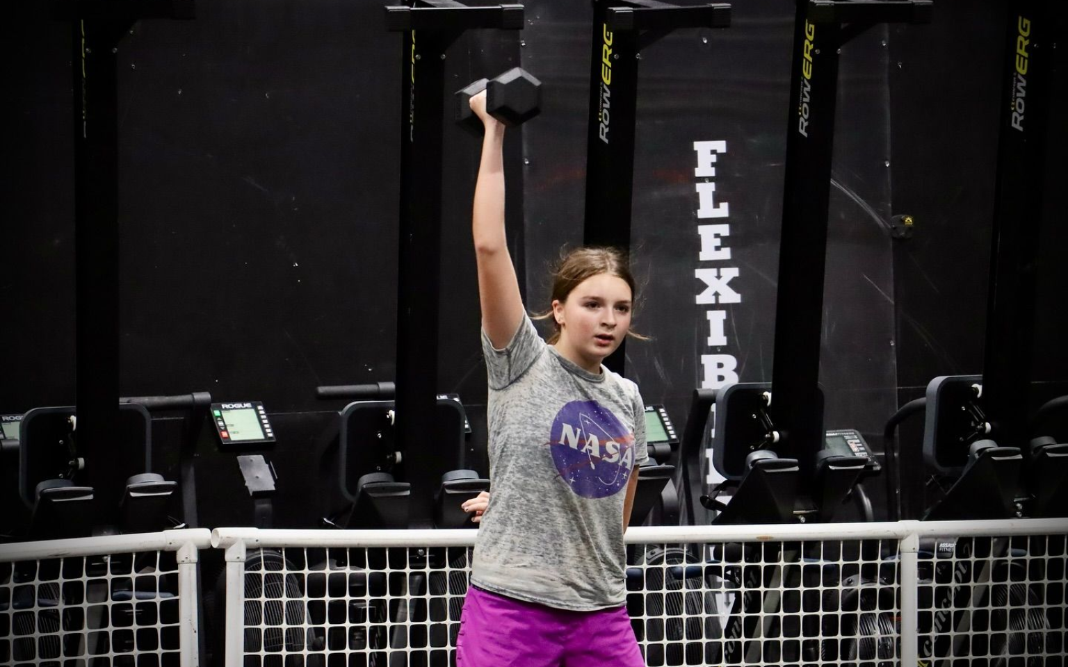 Woman holding a dumbbell overhead in a gym, wearing a gray NASA t-shirt and purple shorts.