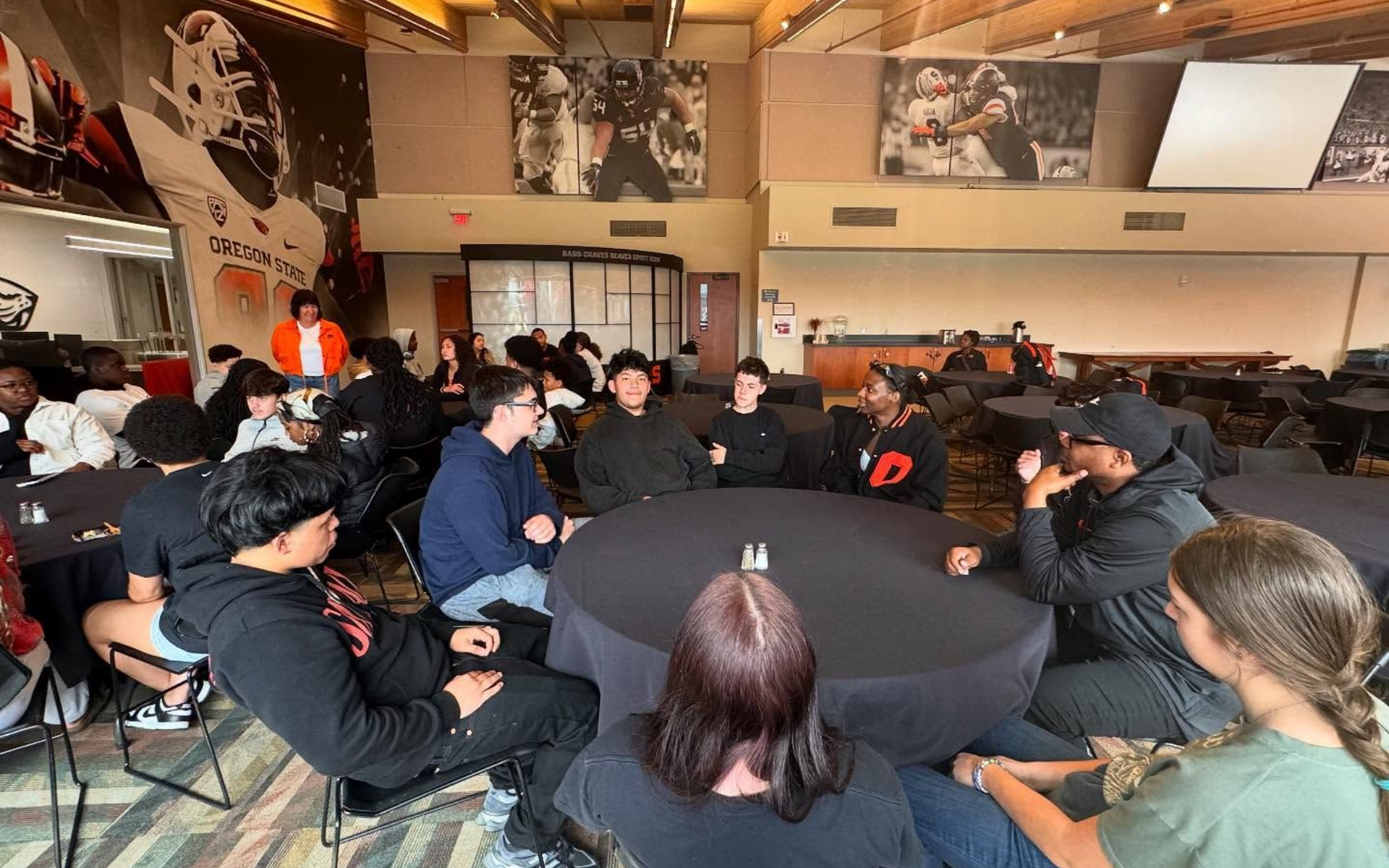 Group of people sitting around tables, talking in a room with sports-themed decor.