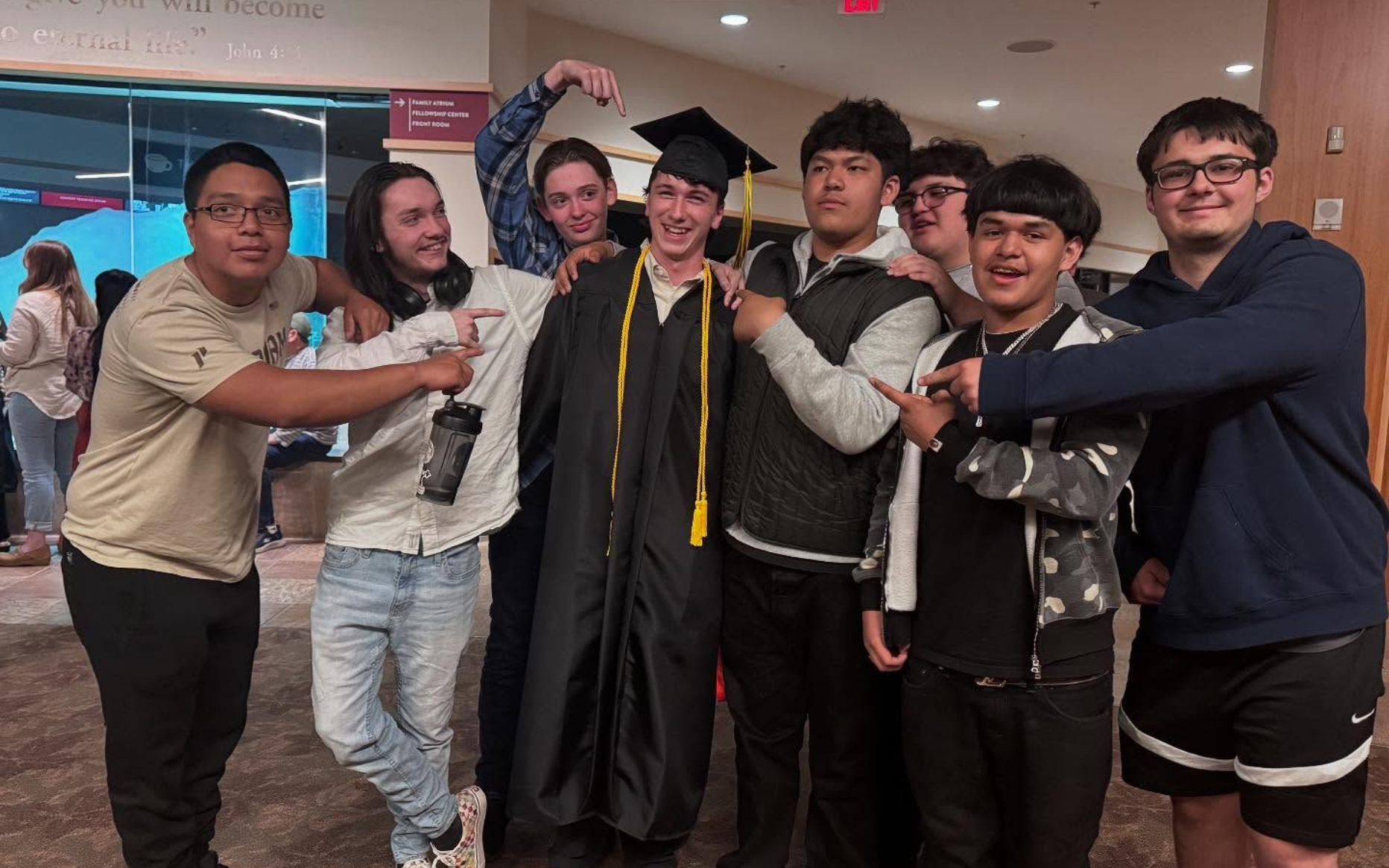Group of young men celebrating; one in a graduation gown, others pointing and smiling. Inside a building.