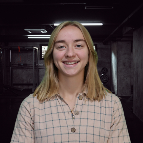 Blonde person with a plaid shirt smiling in a dimly lit room with workout equipment.