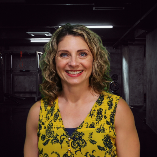 Woman with curly blonde hair smiles, wearing a yellow floral top, against a dark, industrial background.