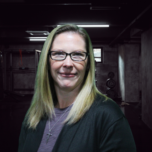 Woman with glasses smiles, in front of a dim basement gym background. She wears a black cardigan and a silver cross necklace.