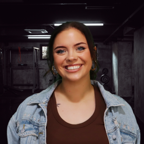 Woman with green-tinted hair and denim jacket smiling in a dimly lit basement with weights.