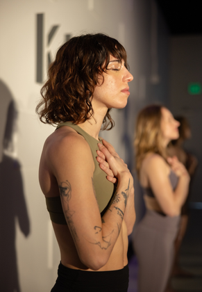 A woman is standing in front of a wall with her eyes closed and hands on her chest.