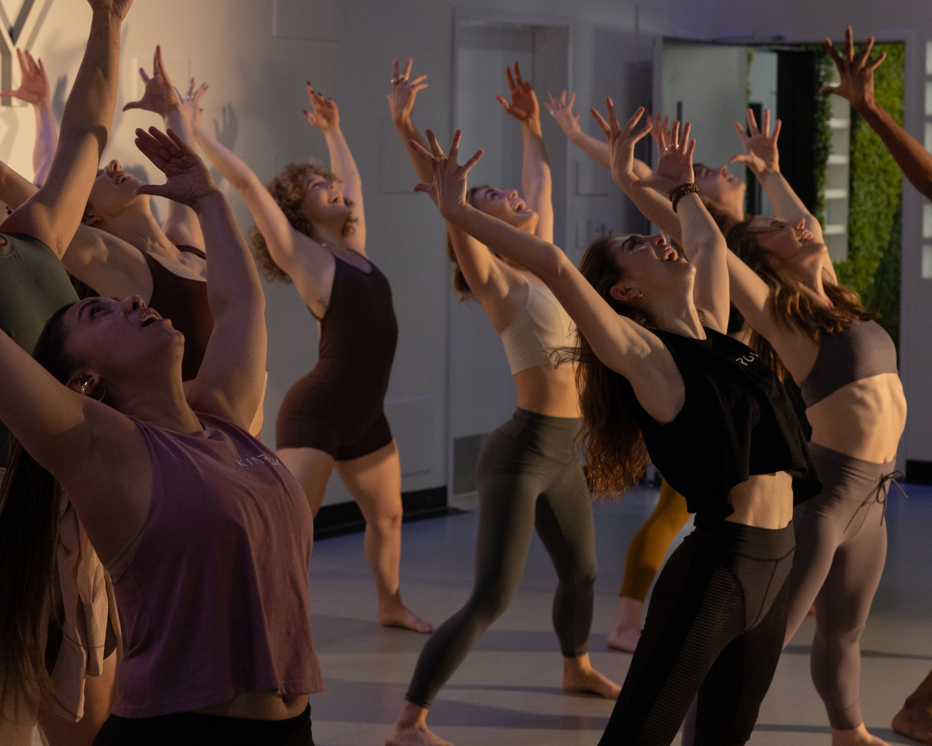 A group of women are dancing in a fitness space with their arms in the air.