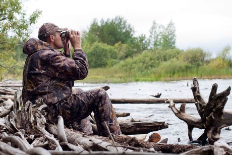 Un homme est assis sur une bûche et regarde à travers des jumelles une rivière.