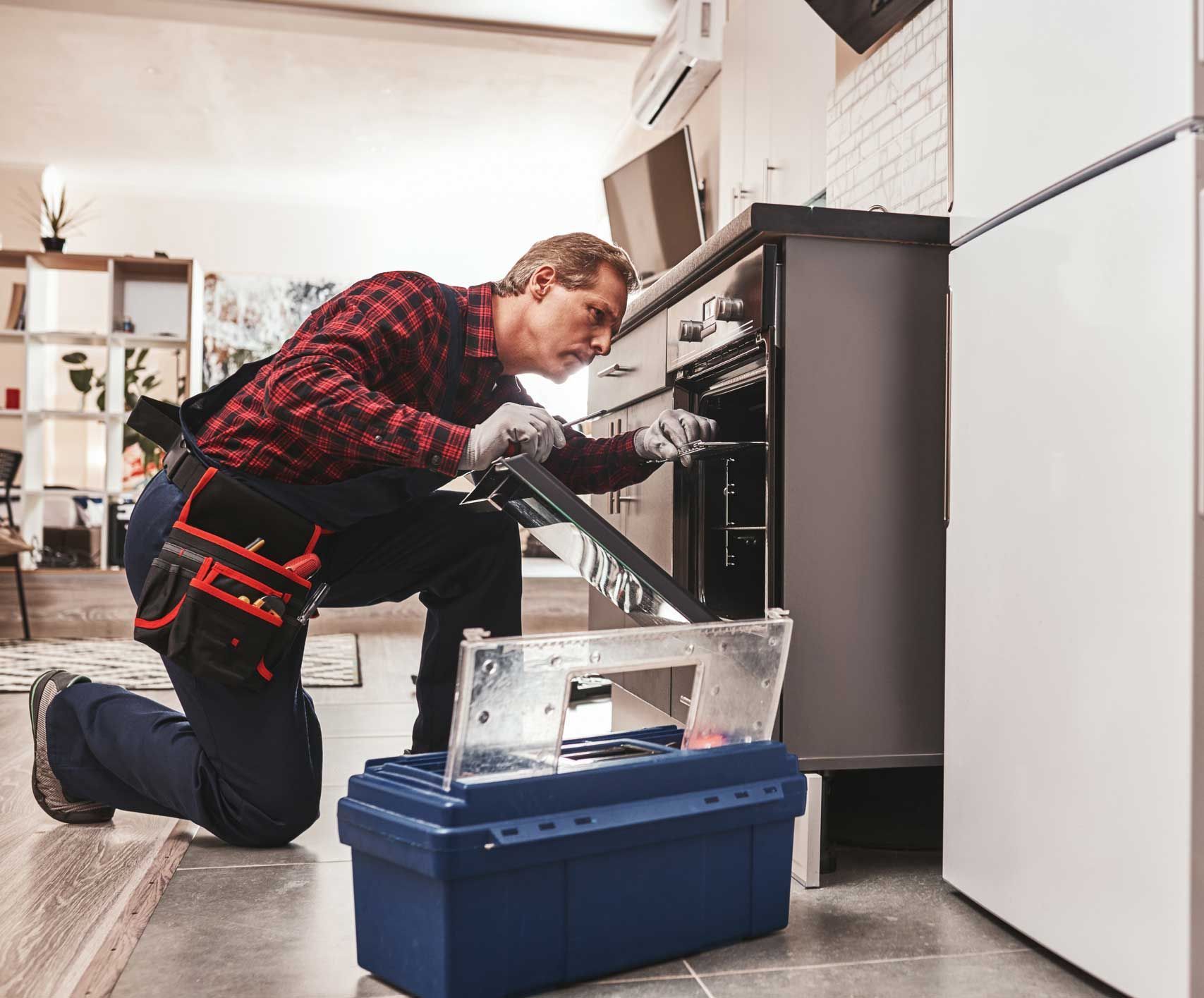 Repairman kneeling, fixing oven, with toolbox. Wearing gloves, red plaid shirt, inside kitchen.