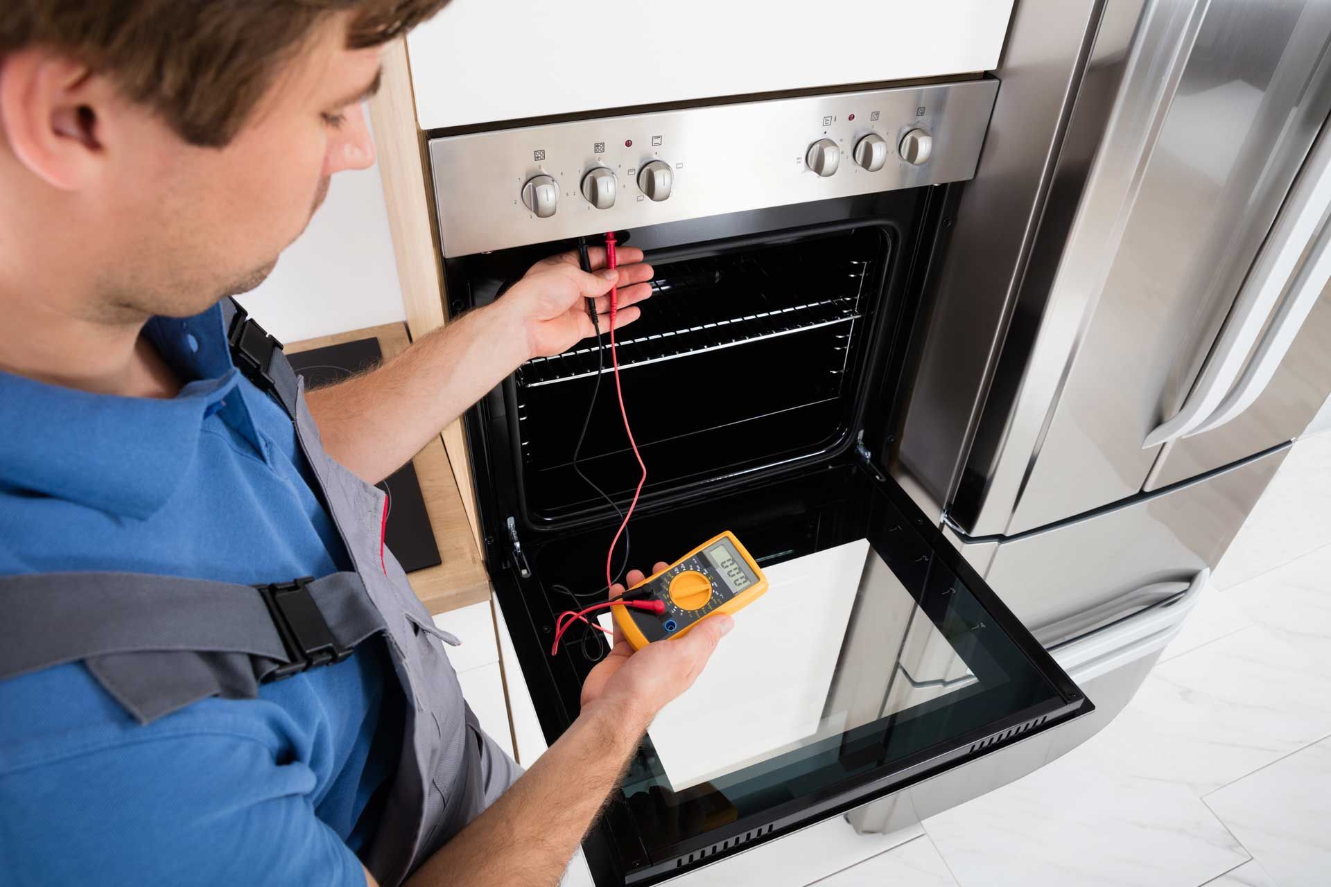 Man testing an oven with a multimeter; oven door open, stainless steel appliances.