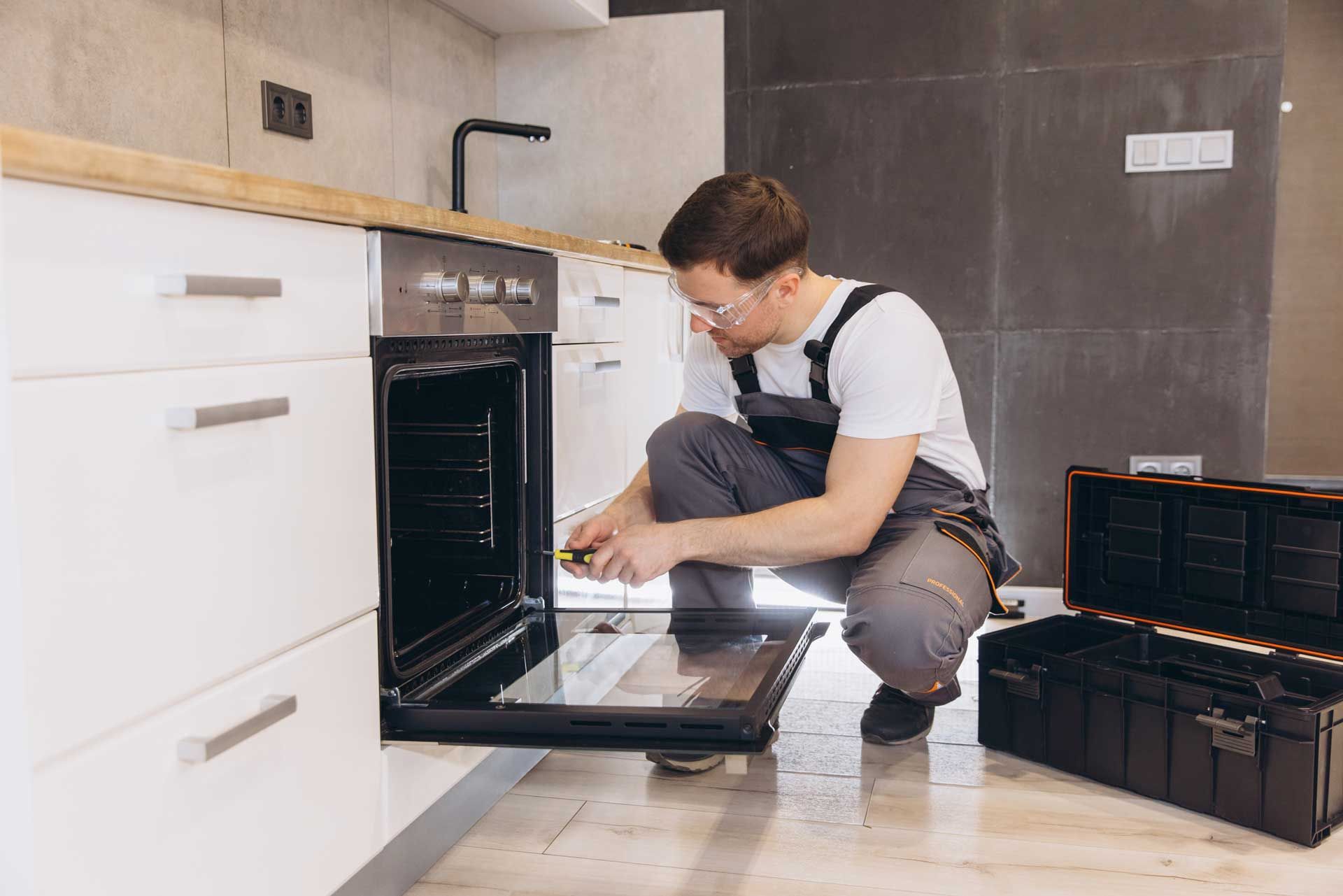 Man in overalls repairs oven in a kitchen, kneeling with a screwdriver. Tool box open nearby.
