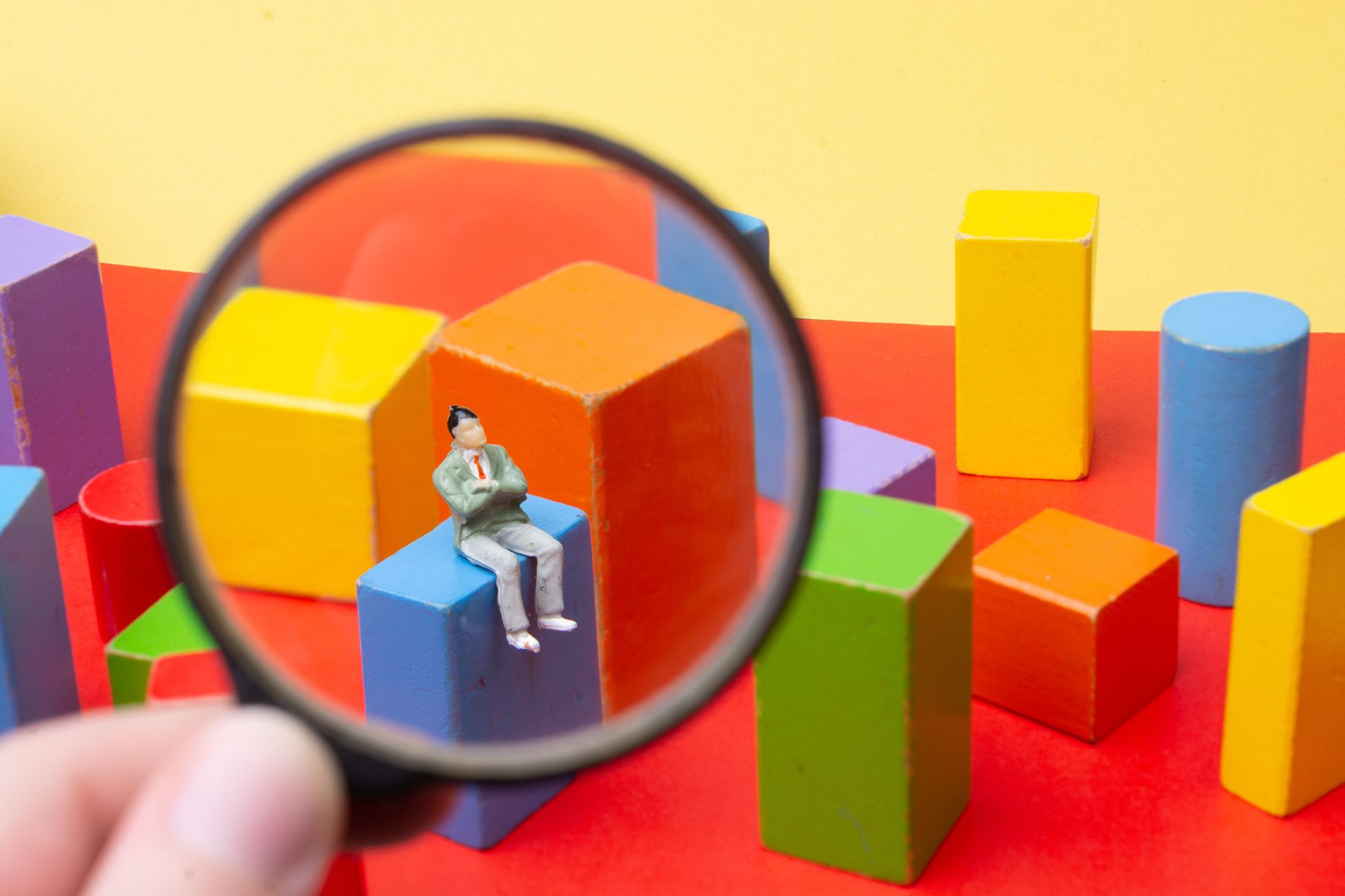 A person is holding a magnifying glass over a miniature man sitting on a block.