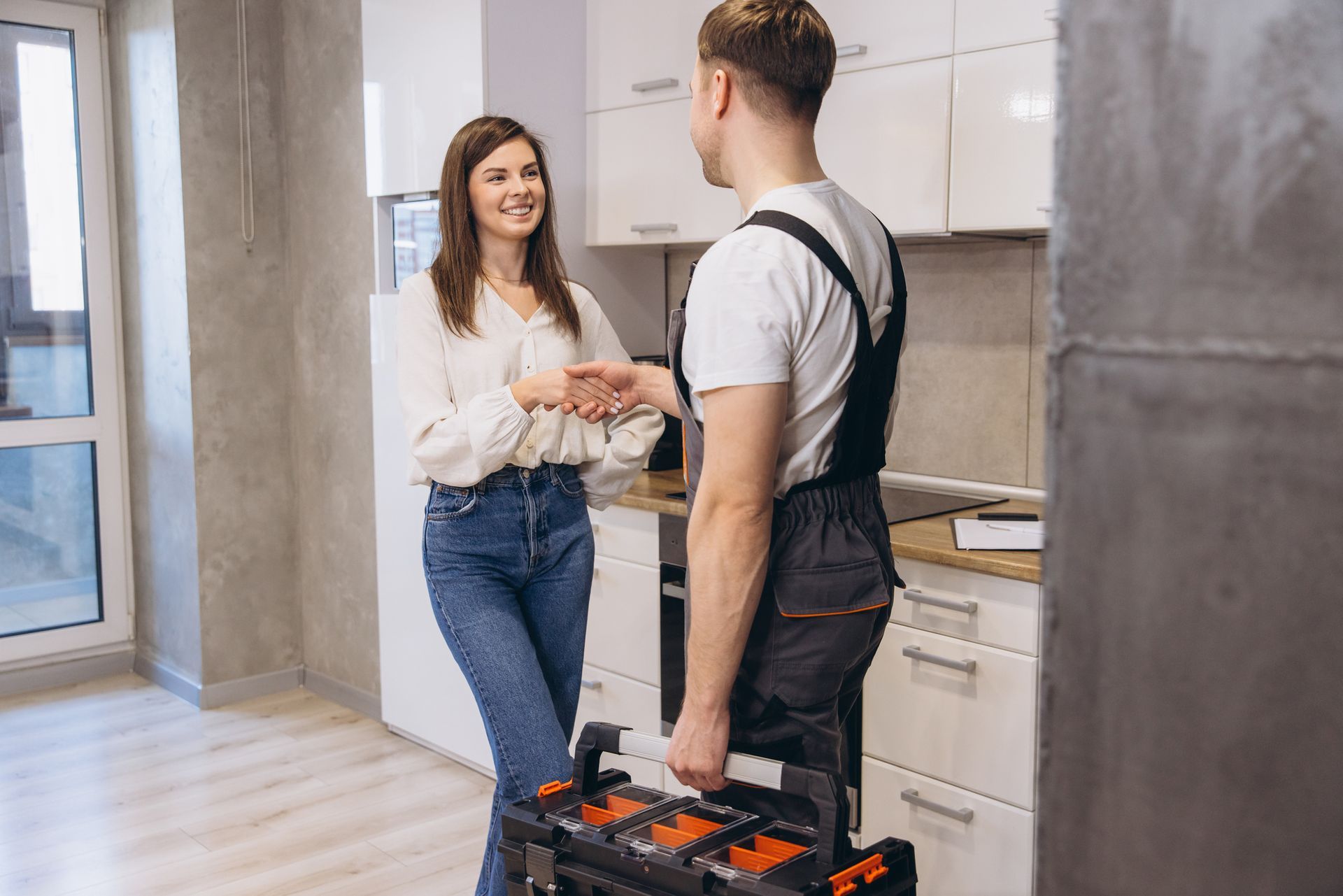 A woman shaking hands with a plumber holding a toolbox in a modern kitchen.