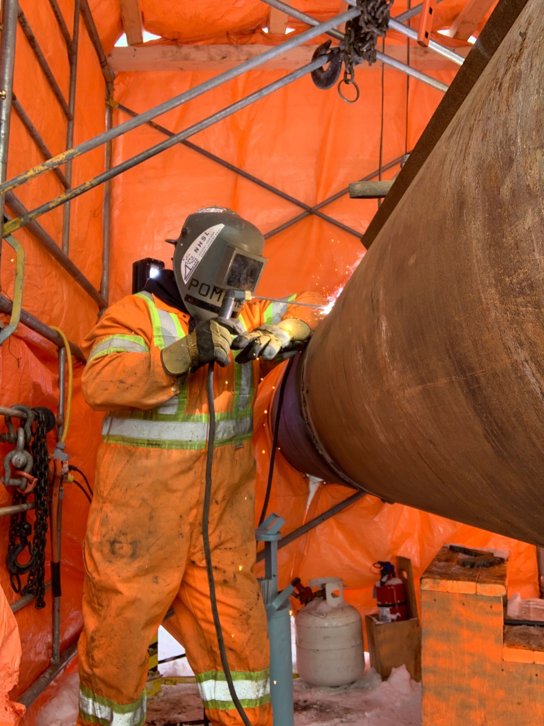 Soudeur en combinaison de protection orange en train de souder un gros tuyau à l'intérieur d'un abri orange.