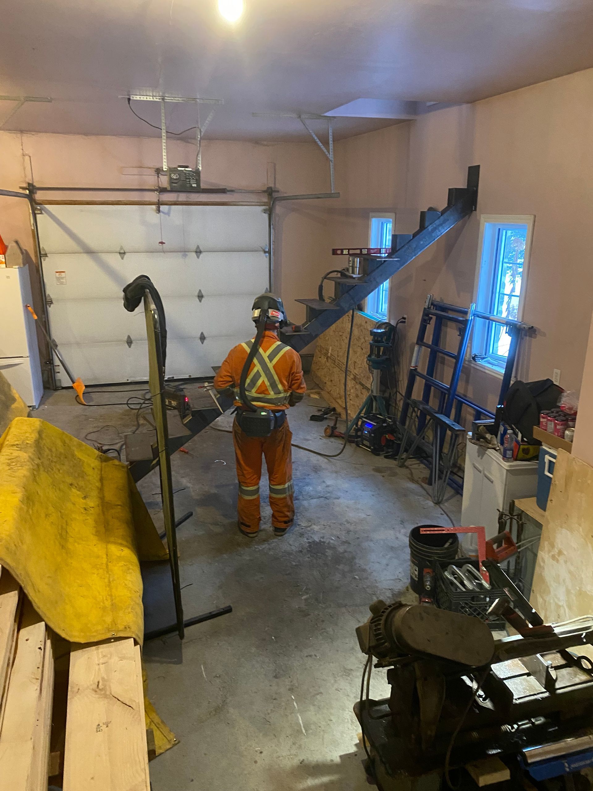 Personne en tenue de travail orange dans un garage en désordre, face à une porte de garage fermée.