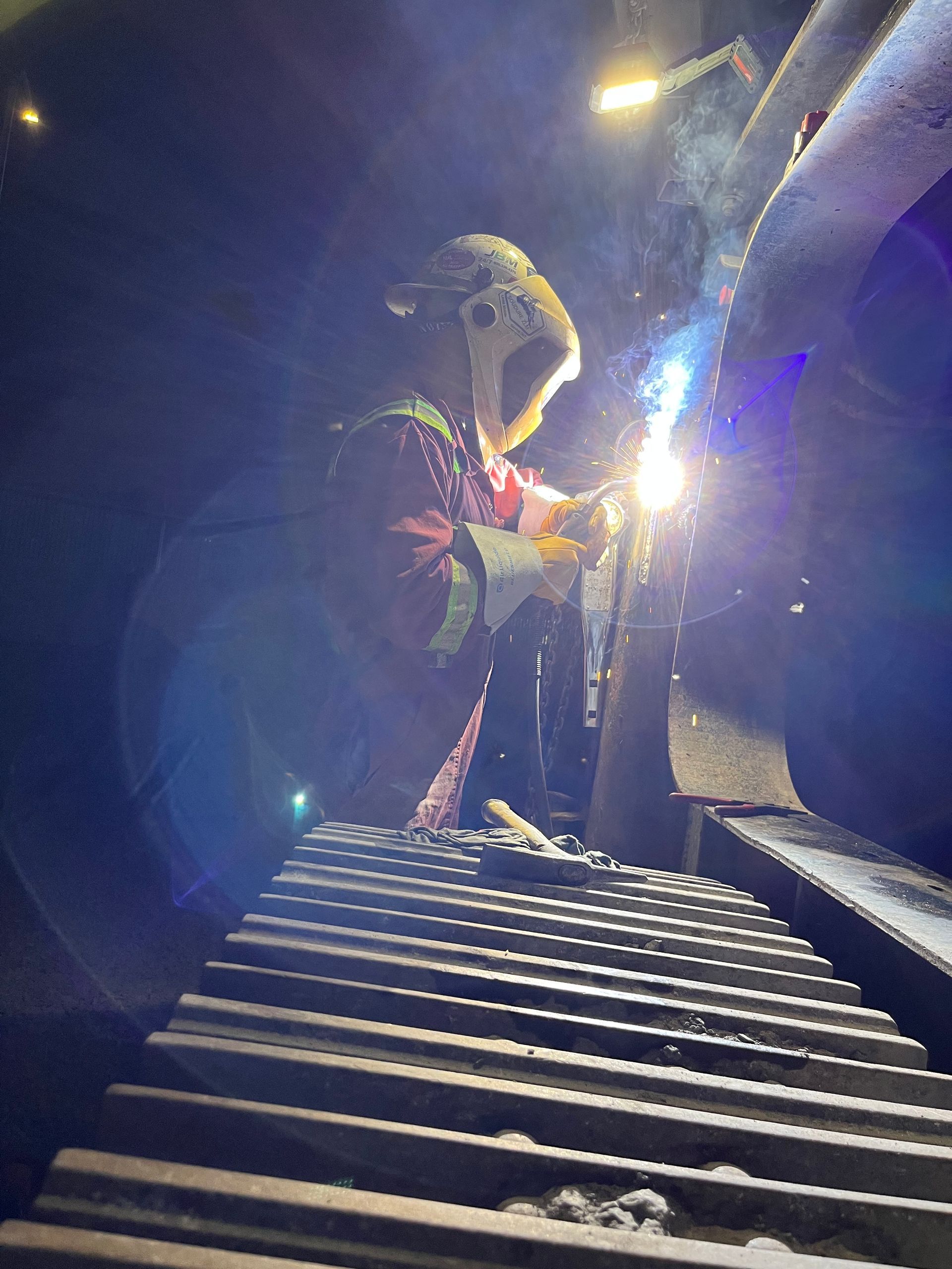 Un soudeur, portant un masque et un équipement de protection, soude du métal dans un environnement industriel faiblement éclairé. Des étincelles et de la lumière sont visibles.