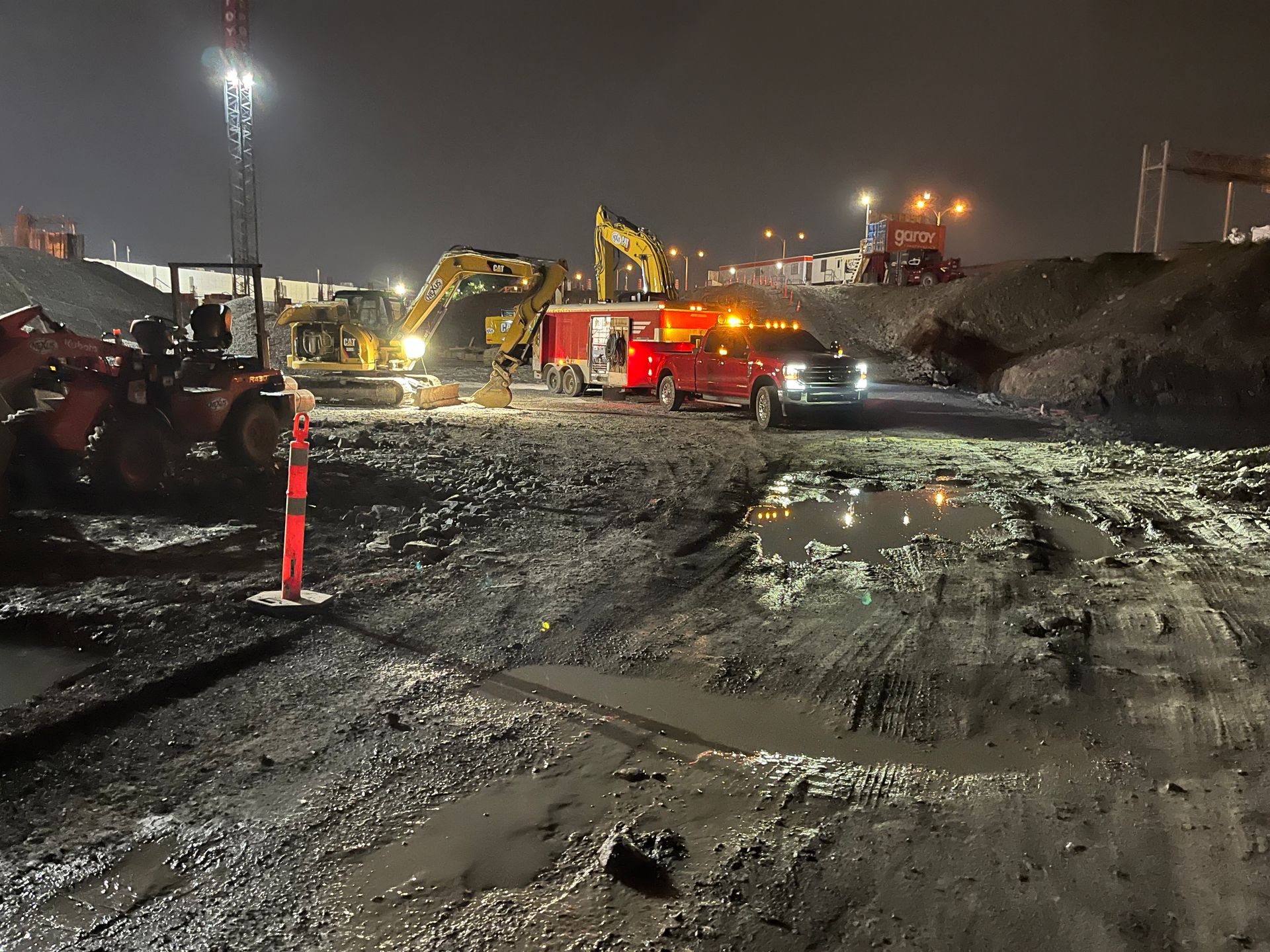 Chantier de construction nocturne avec engins de chantier et camion de pompiers. Sol sombre et boueux.
