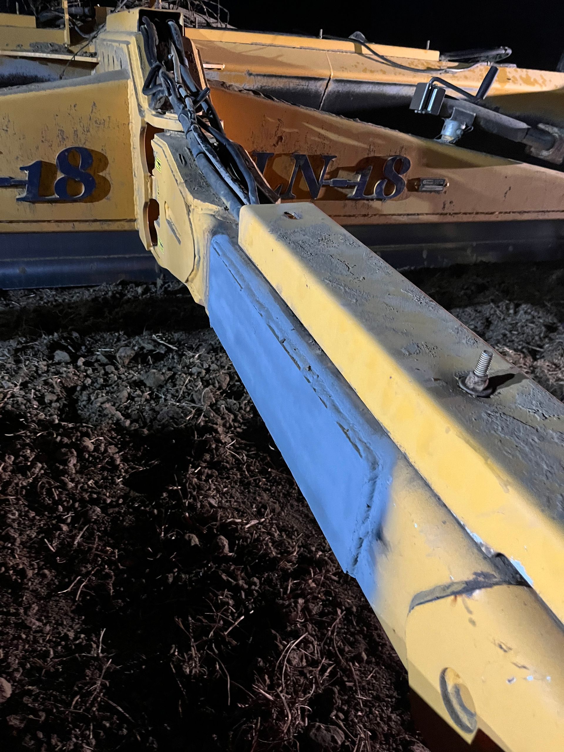 Outil agricole jaune et bleu sur sol sombre, partiellement éclairé la nuit.