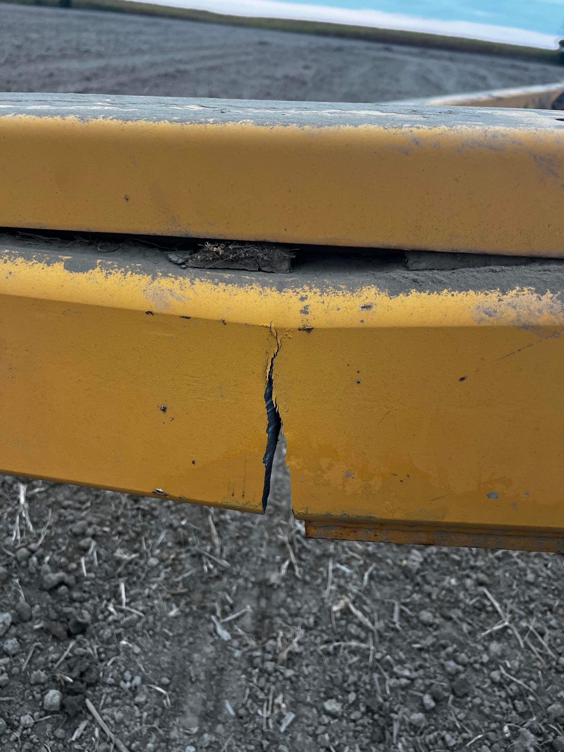 Gros plan sur une poutre métallique jaune présentant une fissure verticale proéminente, posée à l'extérieur sur du gravier.