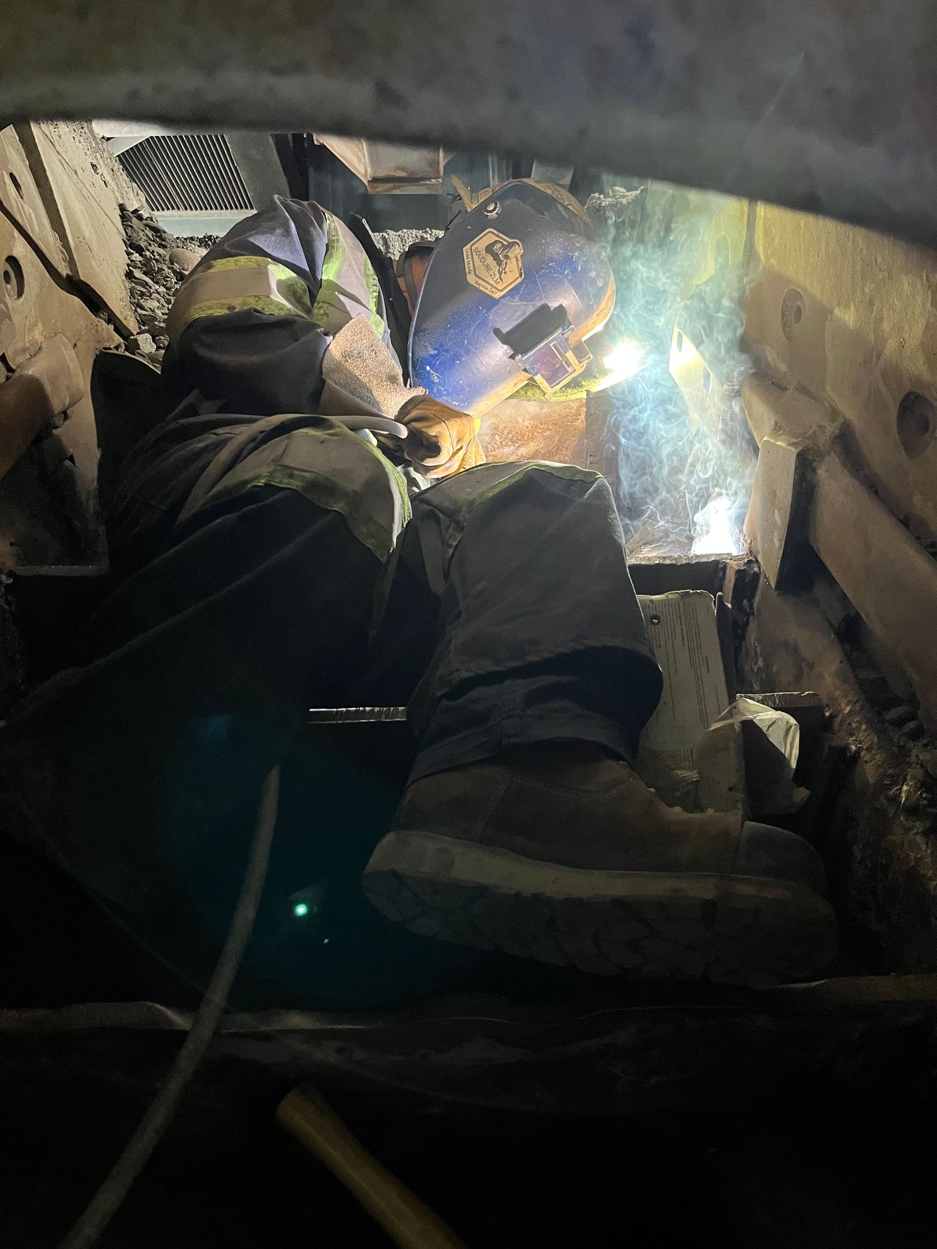 Un soudeur, casqué et botté, travaille dans un espace clos et sombre. Des étincelles jaillissent.