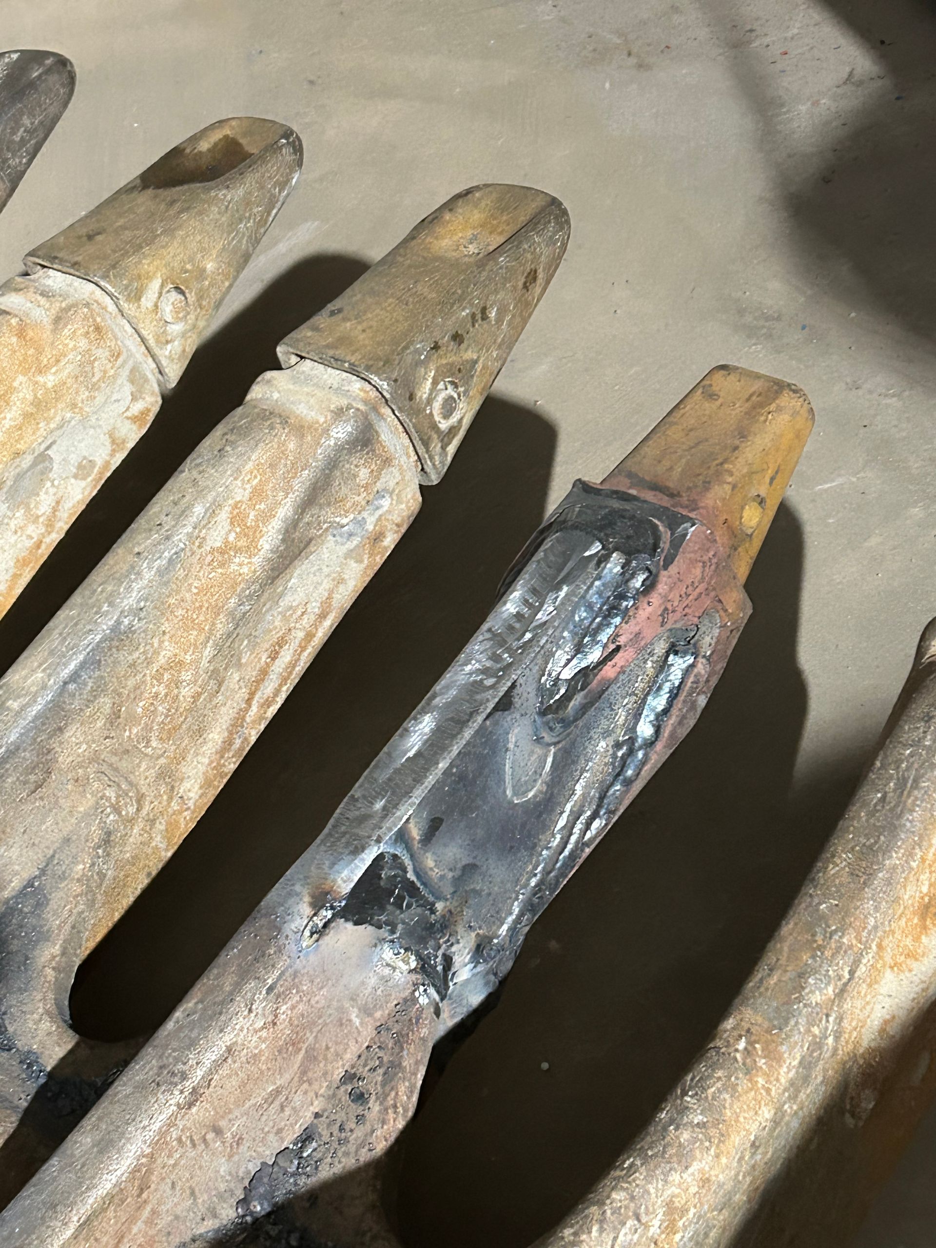 Gros plan sur plusieurs dents métalliques usées d'une excavatrice. Soudure visible sur l'une d'elles.