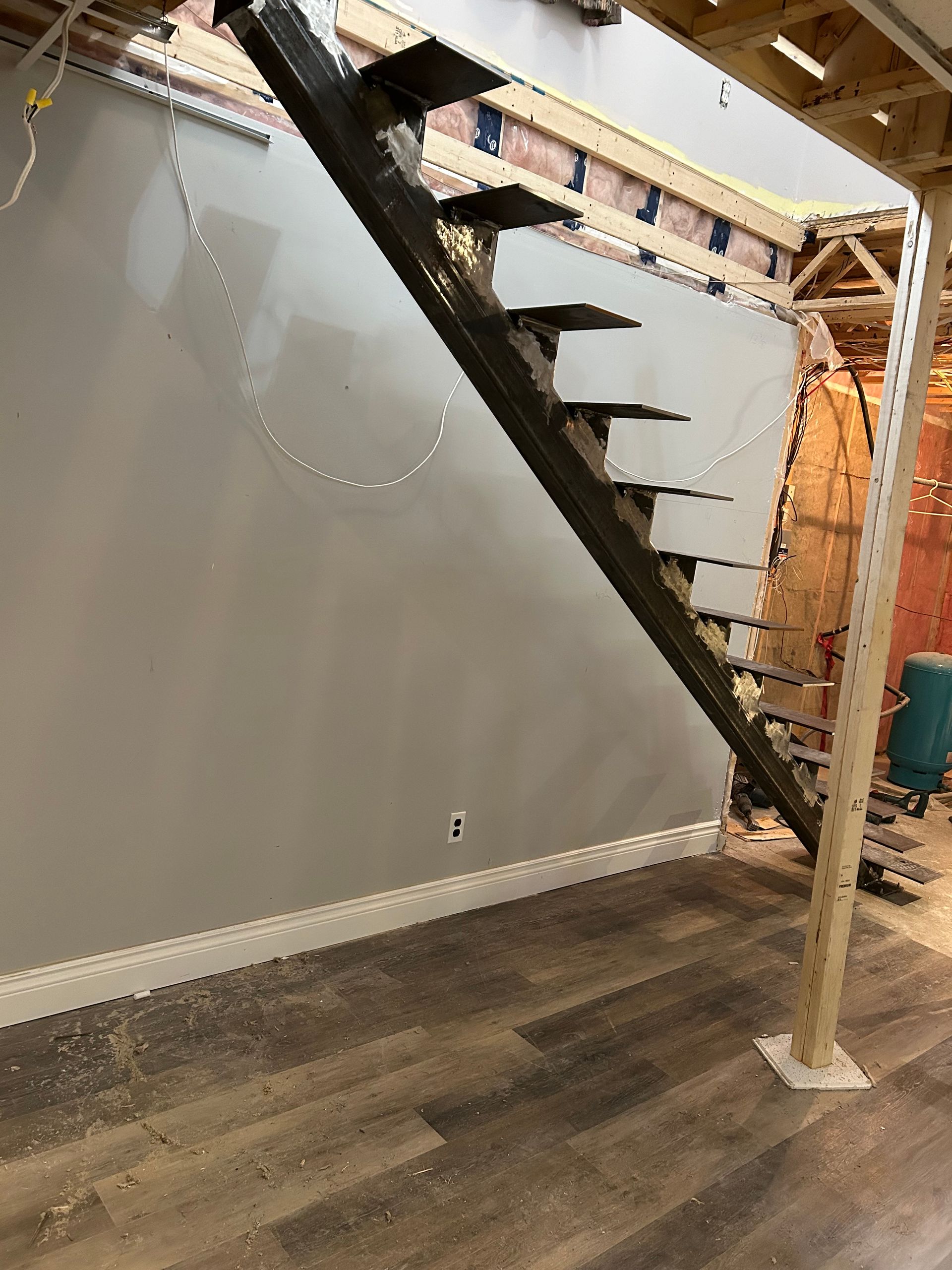 Escalier métallique en construction, avec marches en bois, contre un mur gris et un sol en bois.