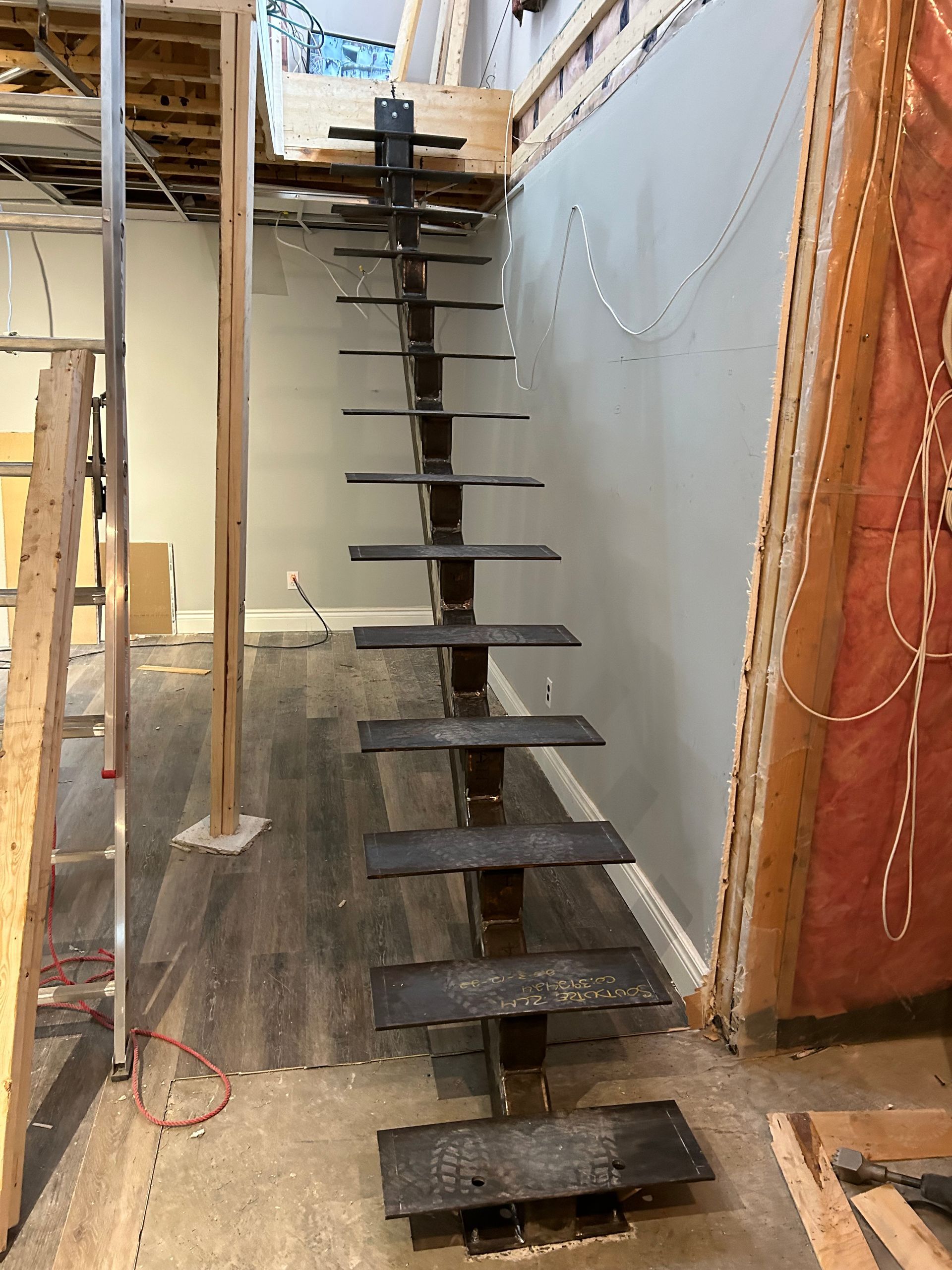 Escalier flottant en métal noir dans un bâtiment en construction. On aperçoit les murs gris, la charpente en bois et le sol.