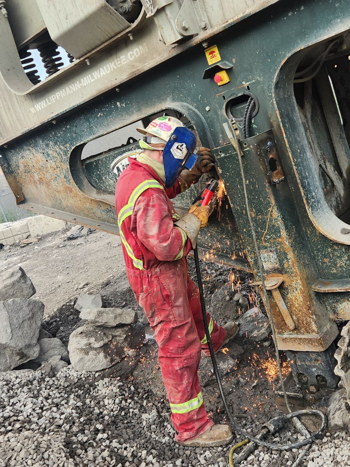 Soudeur en combinaison rouge travaillant sur des machines, des étincelles jaillissent, en extérieur.