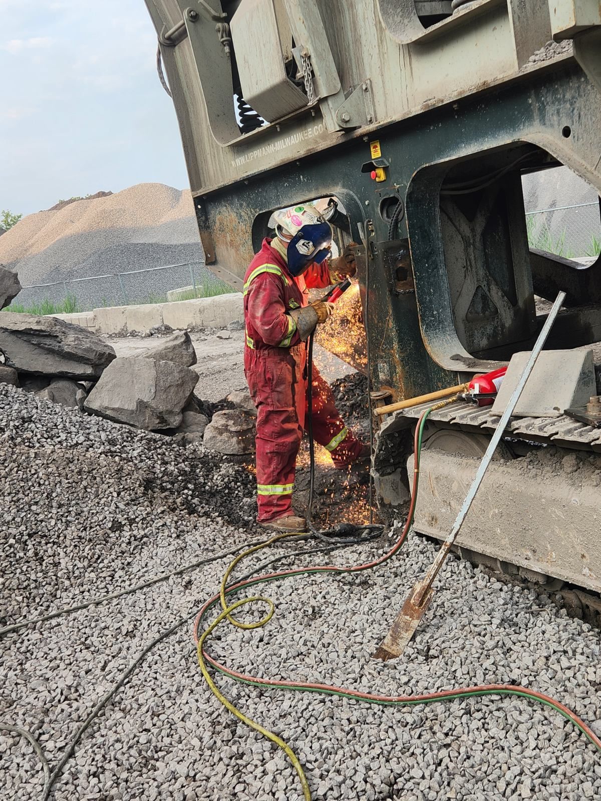 Une personne en tenue de protection soude du métal sur une grande machine industrielle à l'extérieur, des étincelles jaillissent.